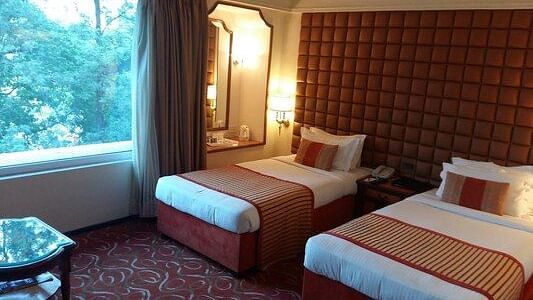 A twin bed setup on carpeted flooring with headboards and ambient lighting, offering a view of the greenery outside. - Pride Hotel Pune