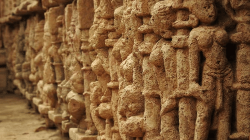 A row of weathered stone sculptures of human figures, closely packed together, lines a textured wall.