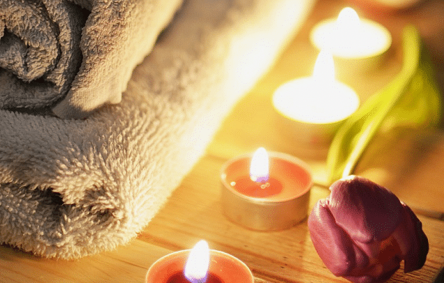 Spa setting with rolled towels, lit candles, flowers, and wooden surface, creating a relaxing atmosphere.