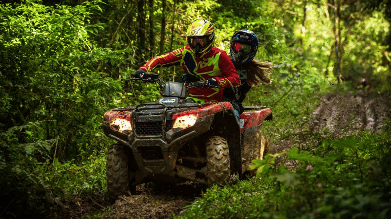 Two people wearing helmets ride a muddy red ATV through a dense green forest trail. The driver is focused while the passenger holds on, as the vehicle moves quickly over rough terrain.