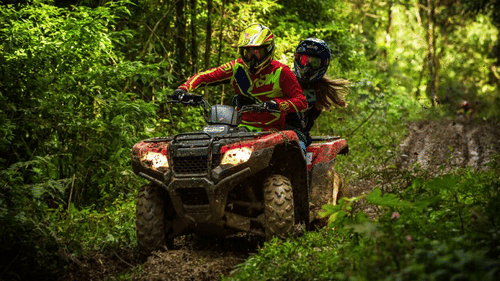 Two people wearing helmets ride a muddy red ATV through a dense green forest trail. The driver is focused while the passenger holds on, as the vehicle moves quickly over rough terrain.