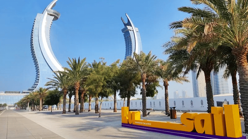 A sunny outdoor scene with palm trees lining a walkway, leading to two tall, curved buildings in the distance. A sign with the word Al Sail is in the foreground.