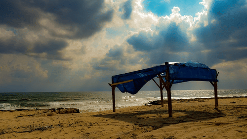 A shelter with a tarp roof set up on sandy beach with waves and clouds in the background.