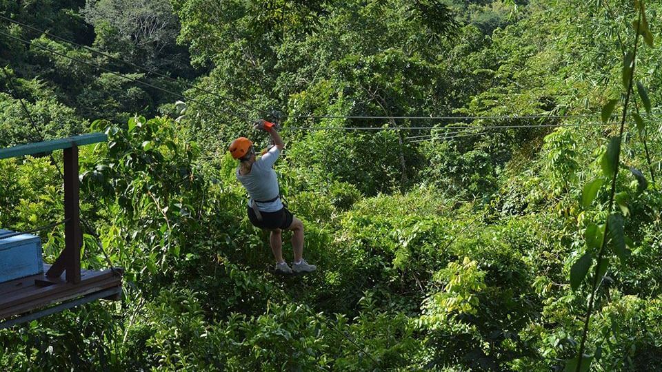 A person wearing safety helmet and safety belts while zip lining, surrounded by lush greenery.