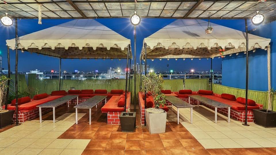 A twilight view of a rooftop dining space with long tables, red cushioned seating and an urban backdrop - TGI Apple Inn, Jaipur 