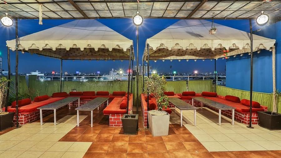 A twilight view of a rooftop dining space with long tables, red cushioned seating and an urban backdrop - TGI Apple Inn, Jaipur 