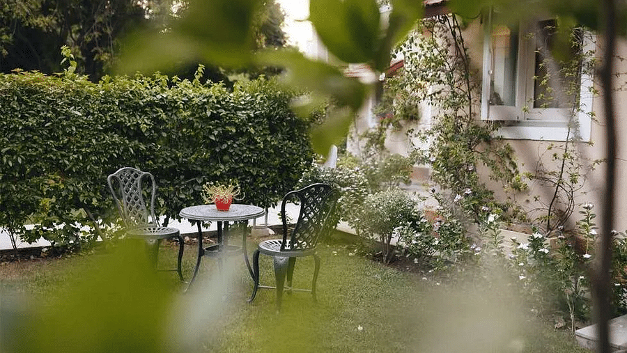 Garden area at Karma Chalets with green hedges, lawn chairs, a table, and flowering plants near the building.