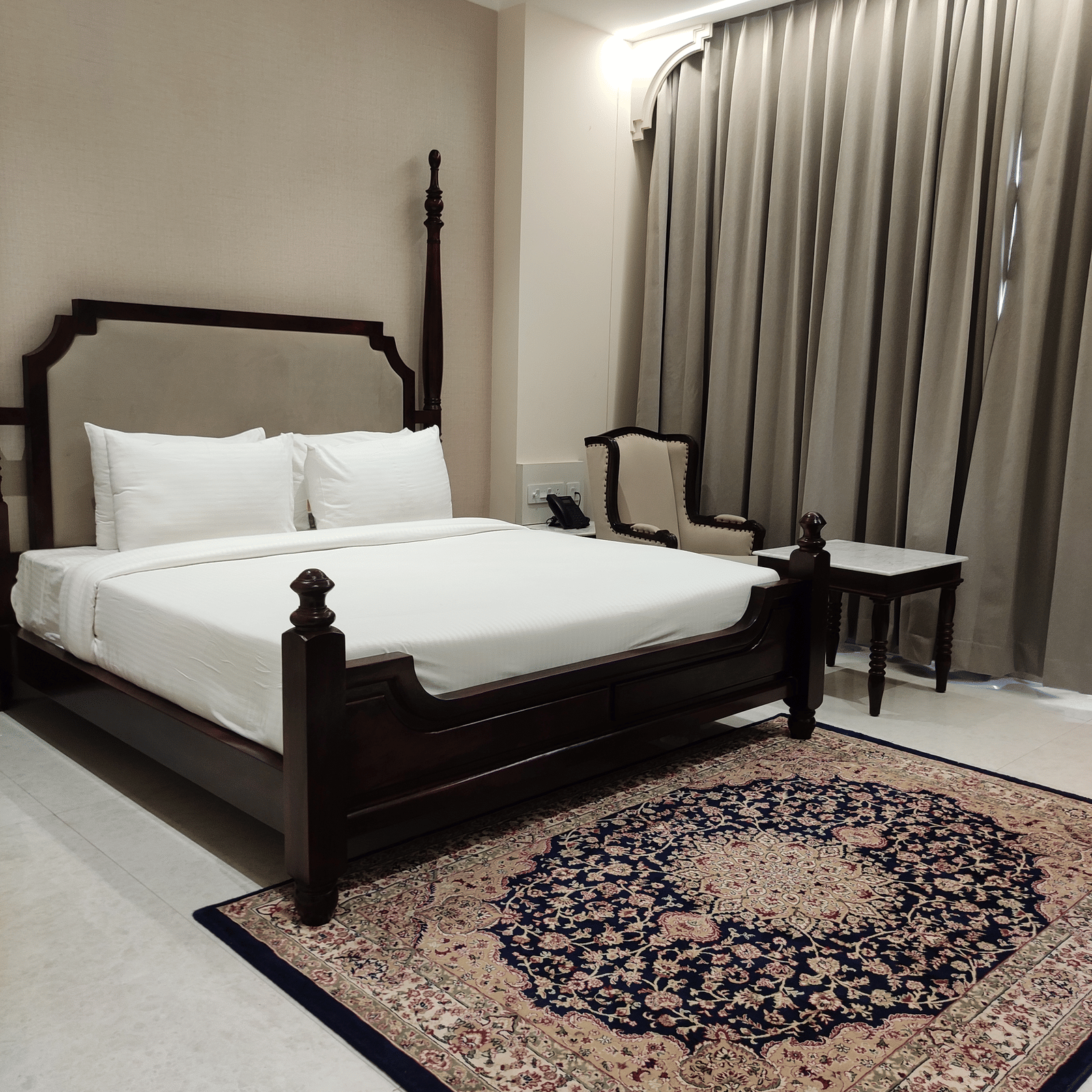 A bedroom at Anant Mahal, Jaipur featuring a king size bed, a patterned rug, floor to ceiling curtains behind a table, and a chair.