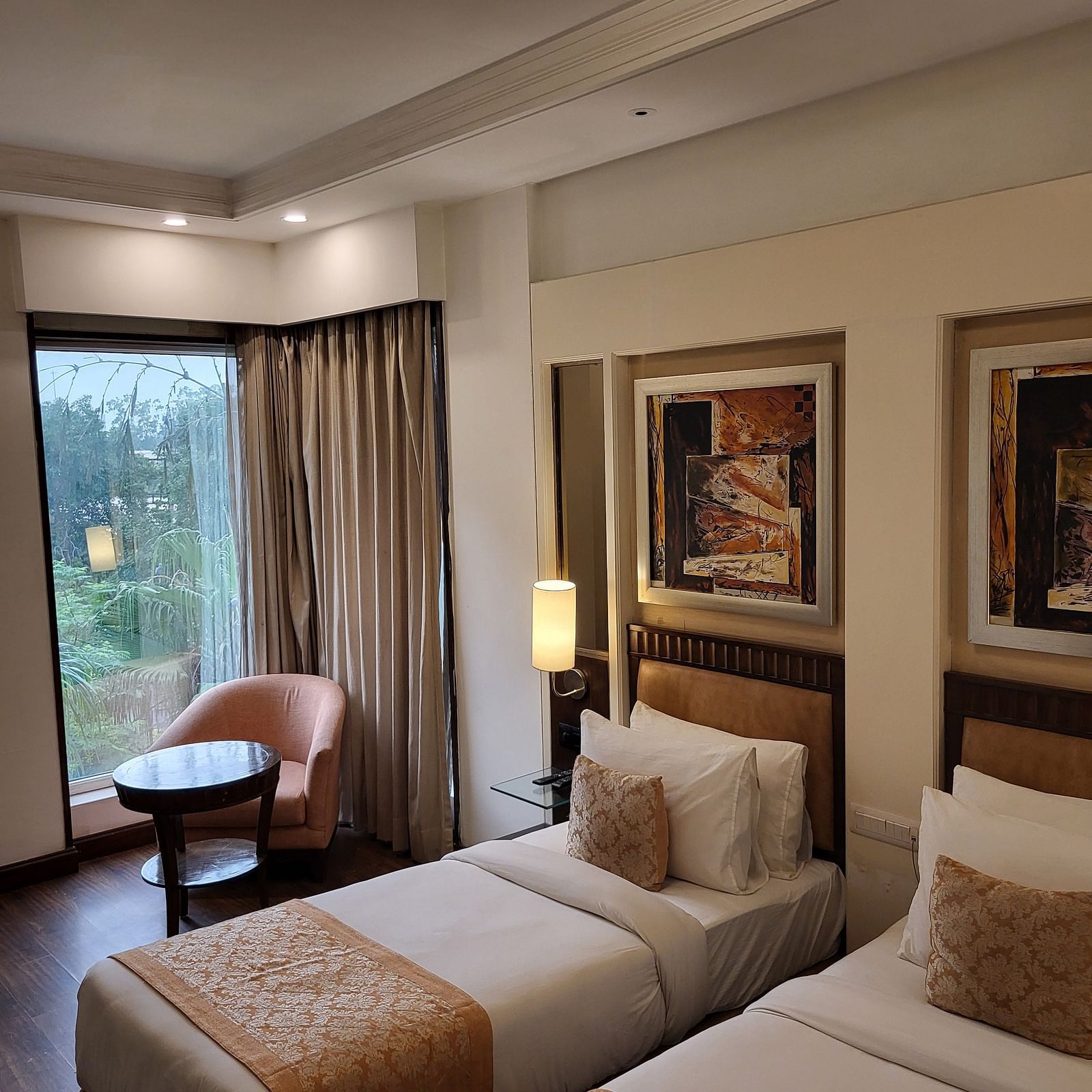 An image of a bedroom featuring twin bed, a sofa chair, a small centre table, bedside table with lamp, photo frames hanged on the wall, and a window o=overlooking the outdoors - Inde Hotel Signature Tower Gurgaon