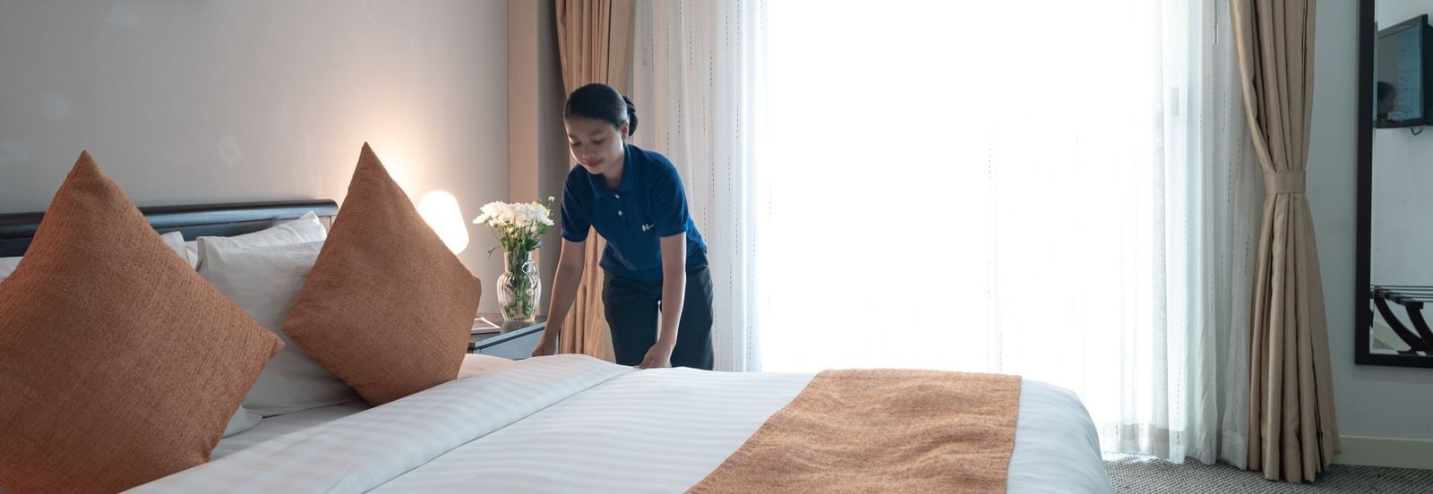 A staff member making a bed, standing beside a large white bed with orange decorative pillows and a matching bed runner near a window with sheer curtains | Central Mansions, Phnom Penh