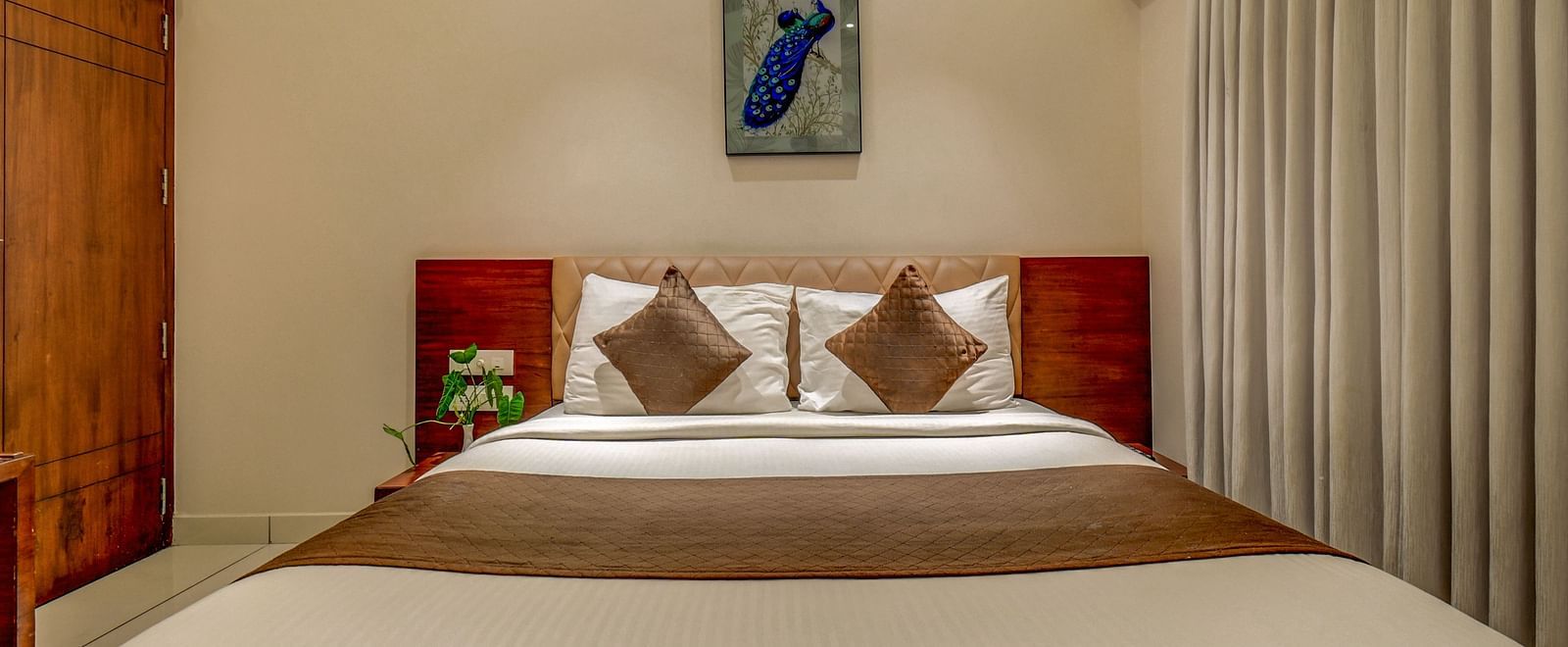 A spacious room at Harigovinda's JNStay with a large bed, wooden headboard, bedside table, curtains pulled to the side, and a photo frame above the headboard