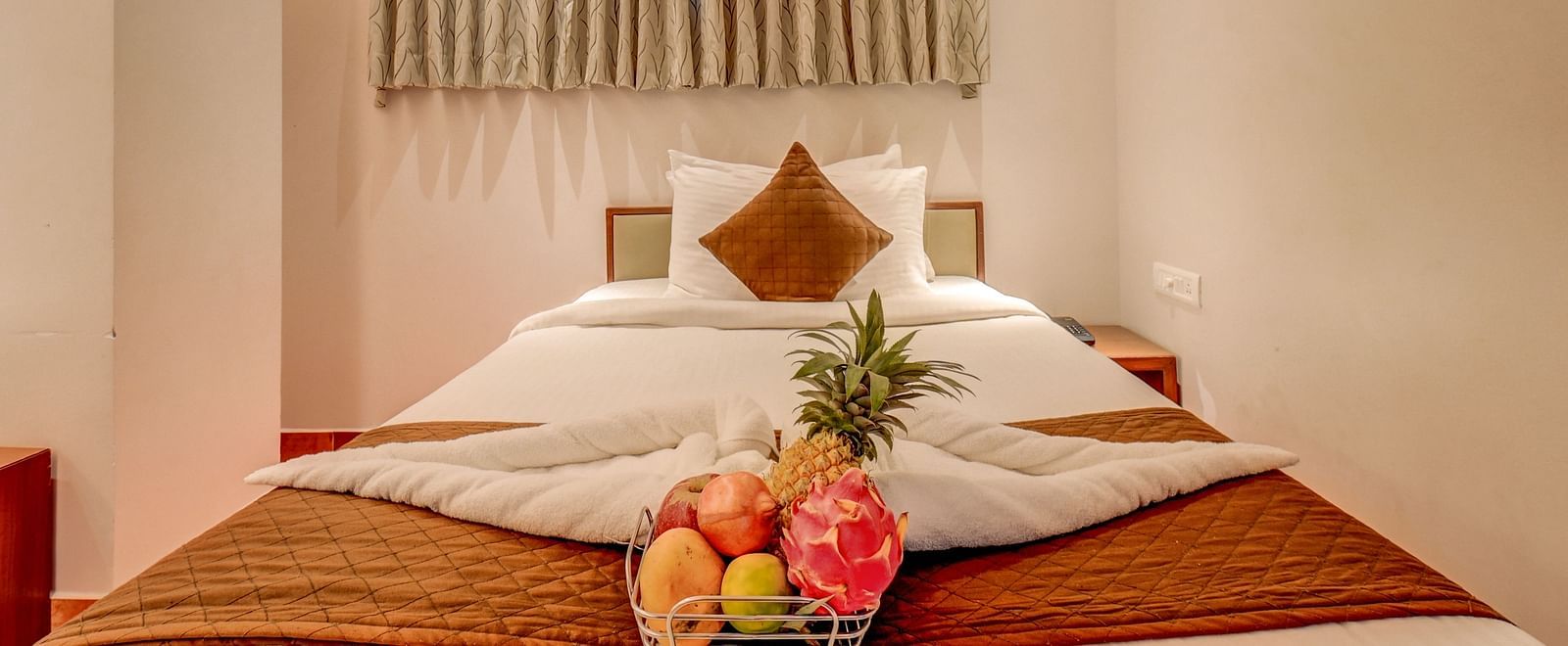 A spacious bed at Harigovinda's JNStay, with neatly arranged blankets, pillows, and a fruit basket, with windows covered by curtains above