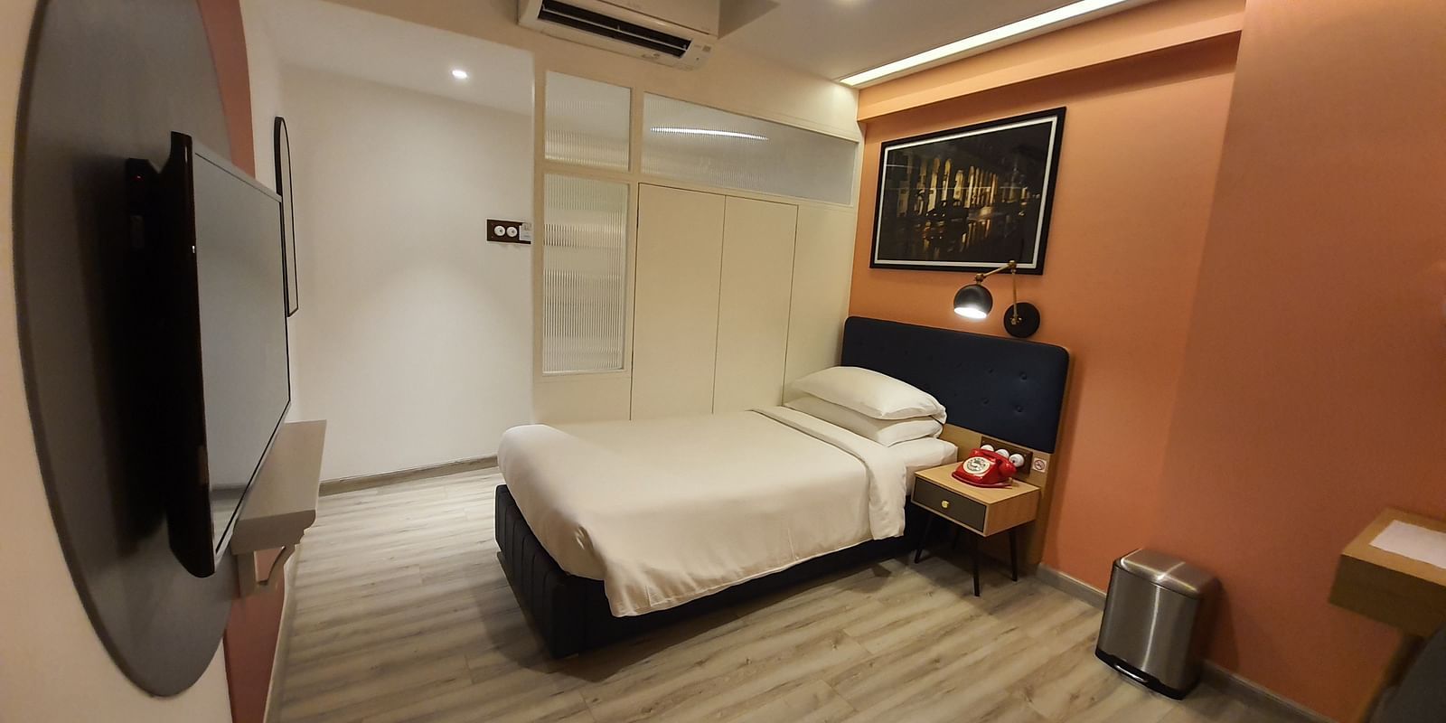 A compact hotel room with a single bed, a small desk, and a flat-screen TV mounted on the wall. The room is decorated in a modern style with orange and white accents.  | T24 Retro
