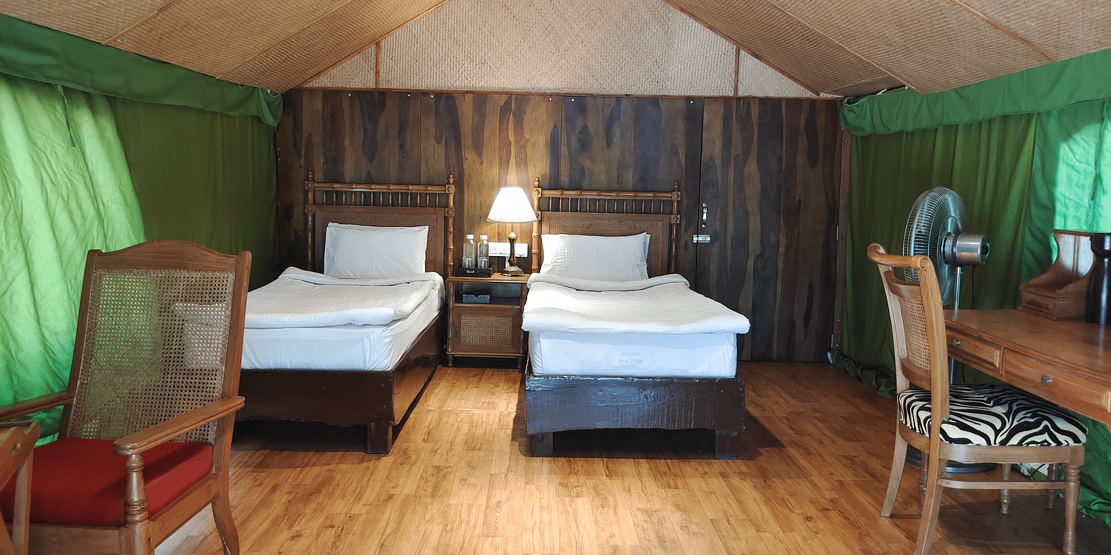 Image featuring Swiss Cottage Tent of The jamoon corbett, with two beds with bed lamp, wooden flooring and a table with two chairs in it.