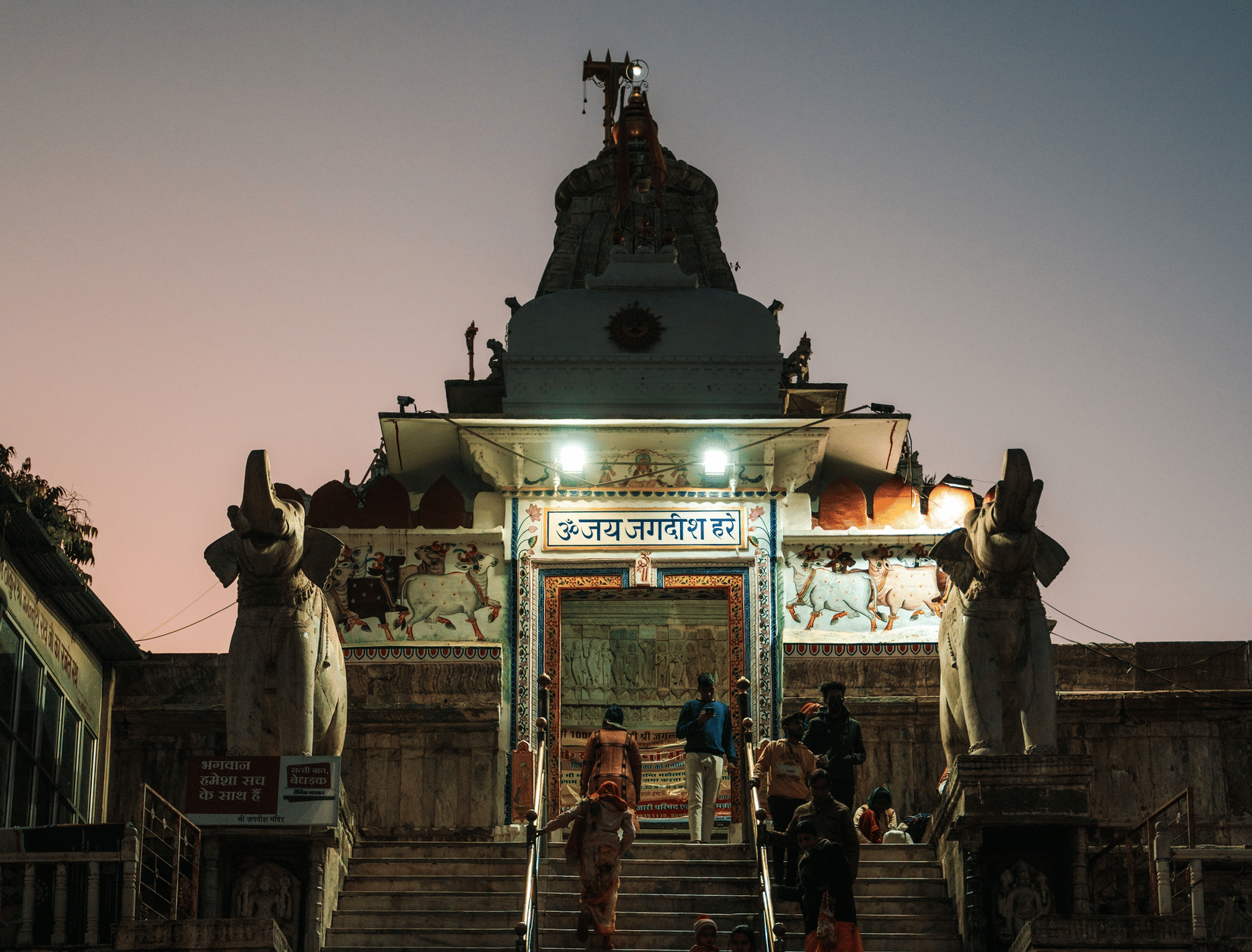 Jagdish Temple Udaipur entrance lit up at dusk with devotees climbing the marble steps between two stone elephant statues