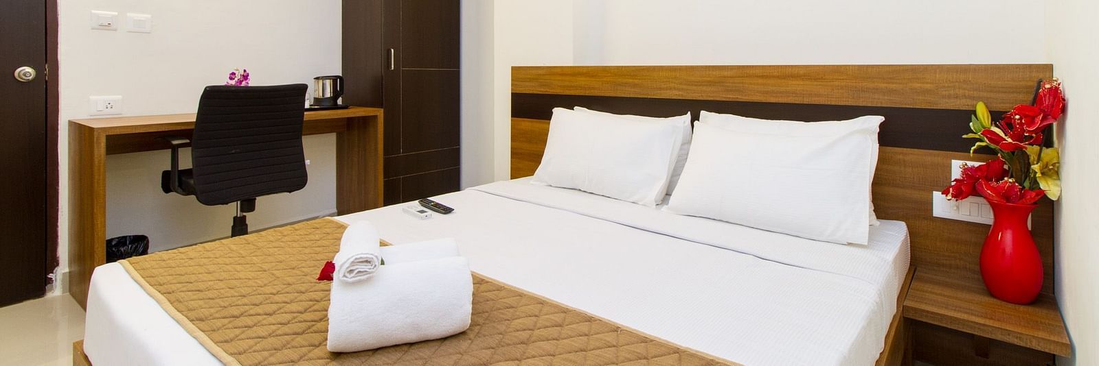 A hotel room including a double bed with brown bedding, a dark wardrobe, and a work desk and chair at Sanctum Suites Whitefield, Bangalore.