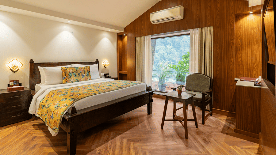 A spacious bedroom featuring a wooden bed, 2 bedside cabinets, a wooden chair facing a small table, and attached balcony with potted plants overlooking the trees outside | Summit by the Ganges Beach Resort & Spa, Rishikesh