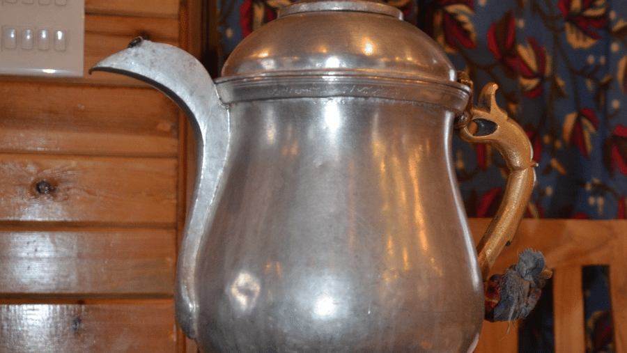 A traditional samovar kettle placed on a patterned surface in front of a wood-panelled wall at Dawat e Drung by FrozenFall Resort, Drung, Tangmarg.
