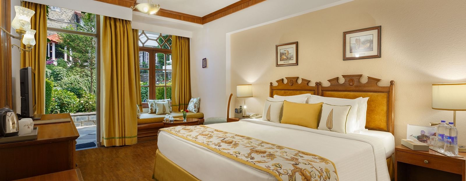 An image of a garden facing room in the naini retreat, nainital, with a king-size bed, wooden furniture, and a window. The room also features a ceiling fan, a TV and wooden flooring.