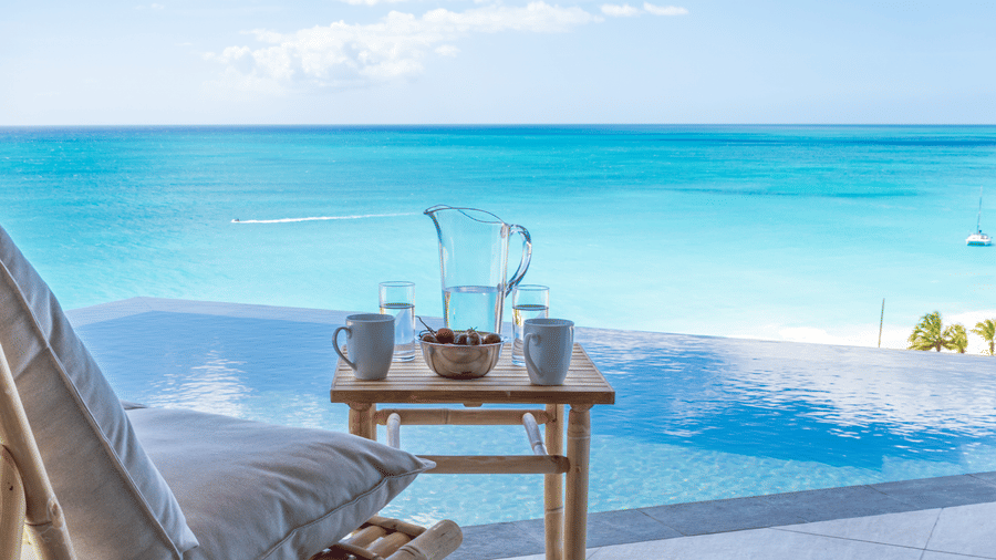 An outdoor setting at the Tamarind Hills Resorts and Villas, Antigua, with two mugs, a bowl of fruit, and a pitcher on a table, with an infinity pool and the sea.