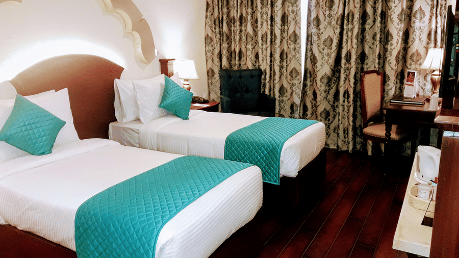 A bedroom at Hotel Sapphire by Eastlynn Collections, Jaipur with 2 single beds, headboard arch design, curtains, armchair, and work desk.