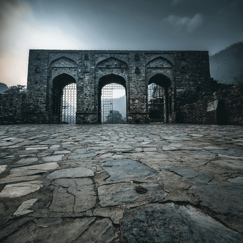 A view of the arch of the Bhangarh Fort with a stone courtyard in front of it. The Bhangarh Fort history is filled with folklore about curses.