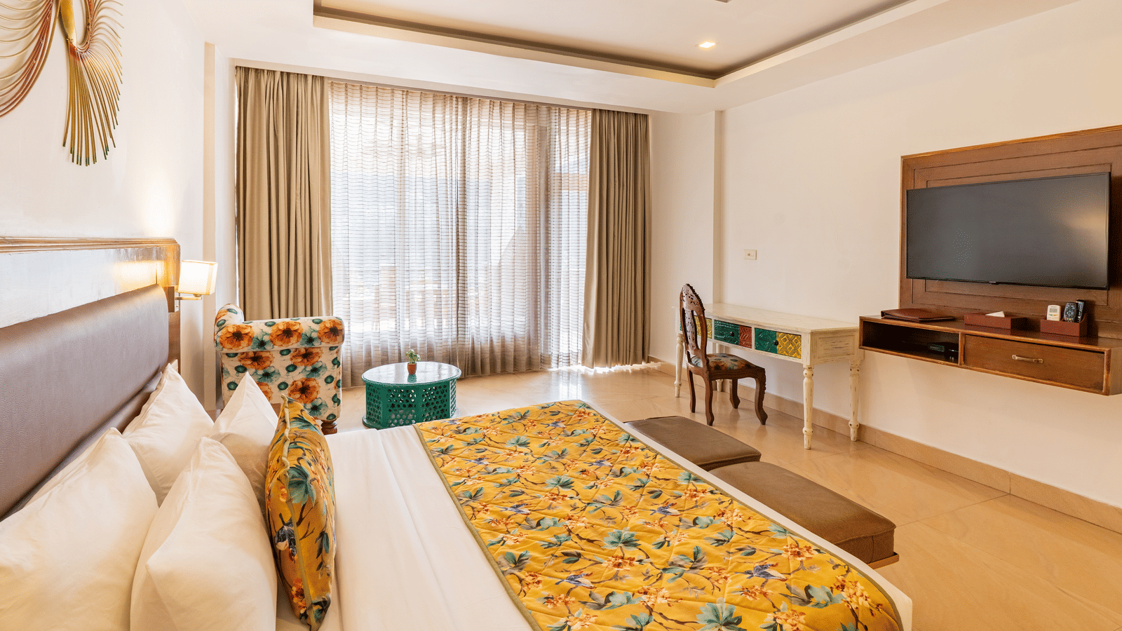 A decorated bedroom featuring a bed, a sofa with a small coffee table, a writing desk with a wooden chair, a television, and curtains drawn | Summit by the Ganges Beach Resort & Spa, Rishikesh 