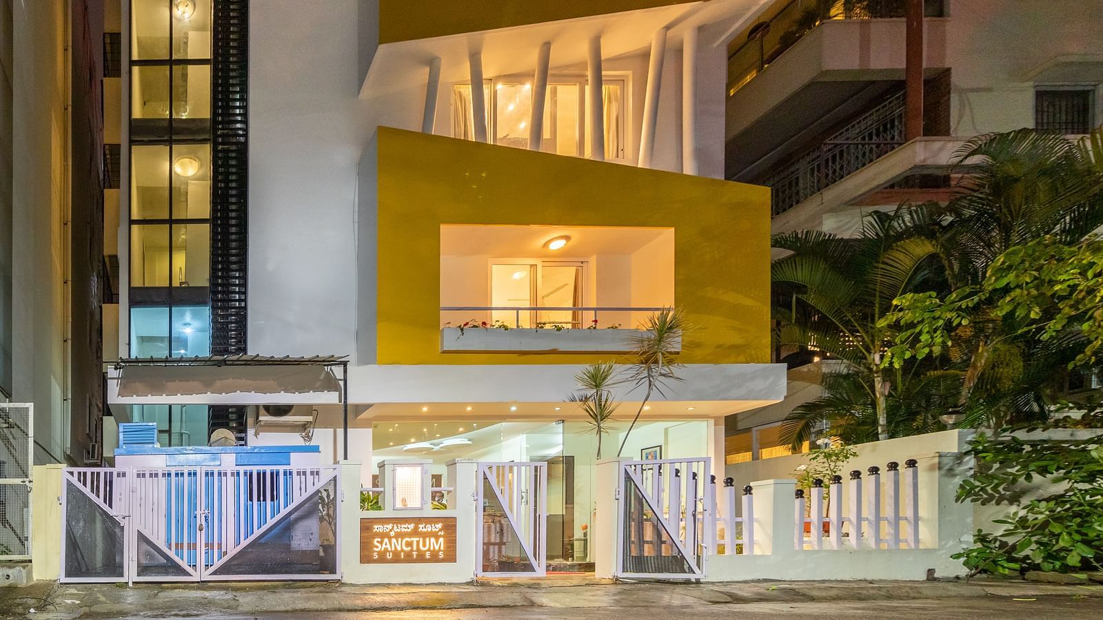 A facade of Sanctum Suites Domlur, Bangalore with warm lighting and view of balconies shot at night.