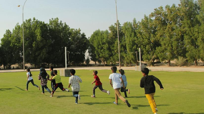A group of children running around the large, open green field under a clear sky.