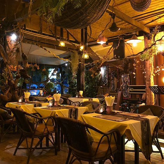 An atmospheric outdoor restaurant or cafe scene at night, with tables and chairs illuminated by warm lights.