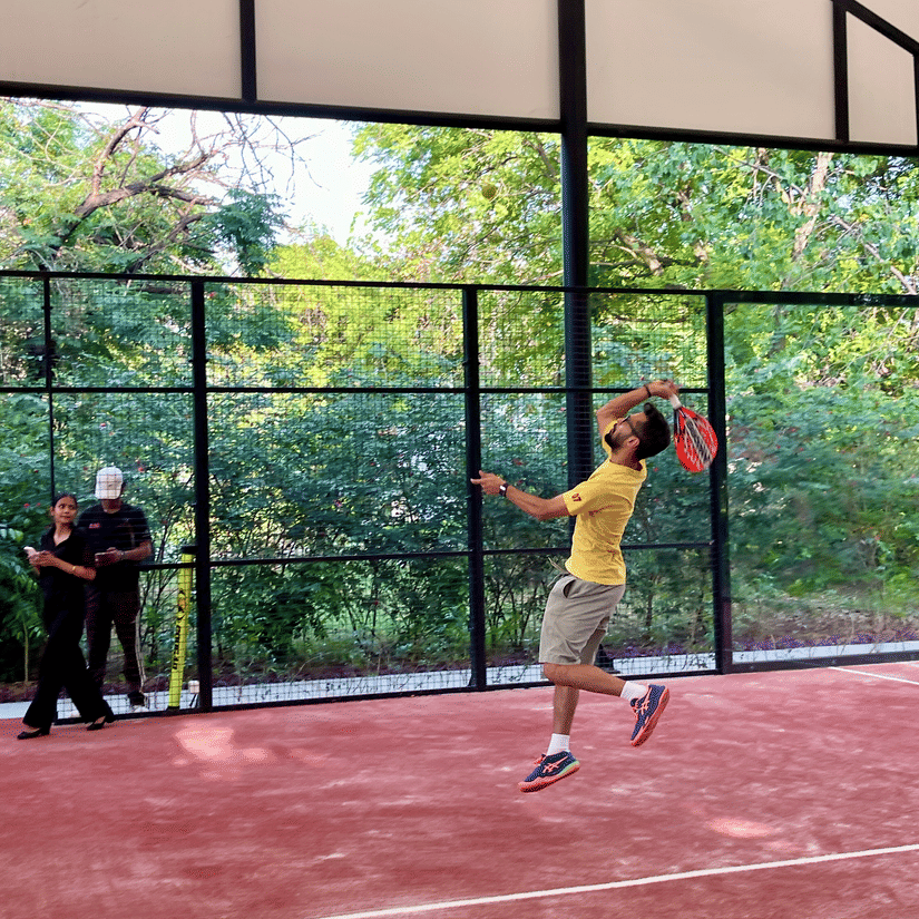 A man in a yellow shirt jumping to hit a ball on a red padel tennis court surrounded by greenery - Karma Lakelands