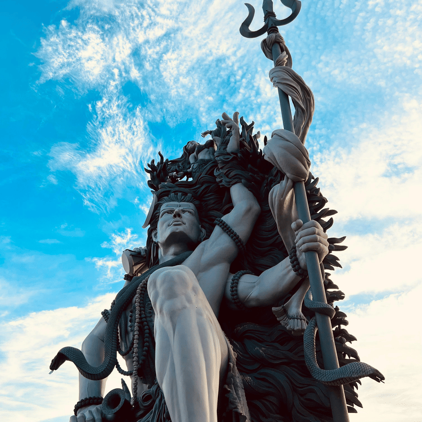 A tall statue of Lord Shiva and a trident, viewed from below against a bright blue sky with scattered clouds.