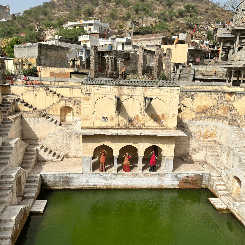 A high-angle view of a stone stepwell containing green water, surrounded by arid hills and local structures.