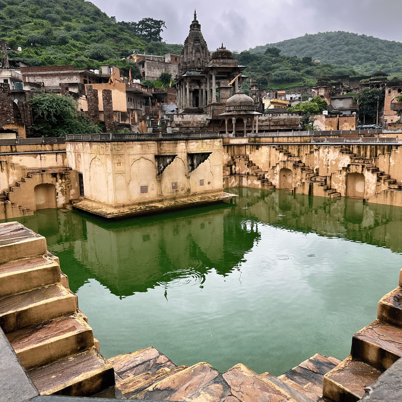A scenic view of a traditional stone stepwell filled with green water, set against hills and old temples.