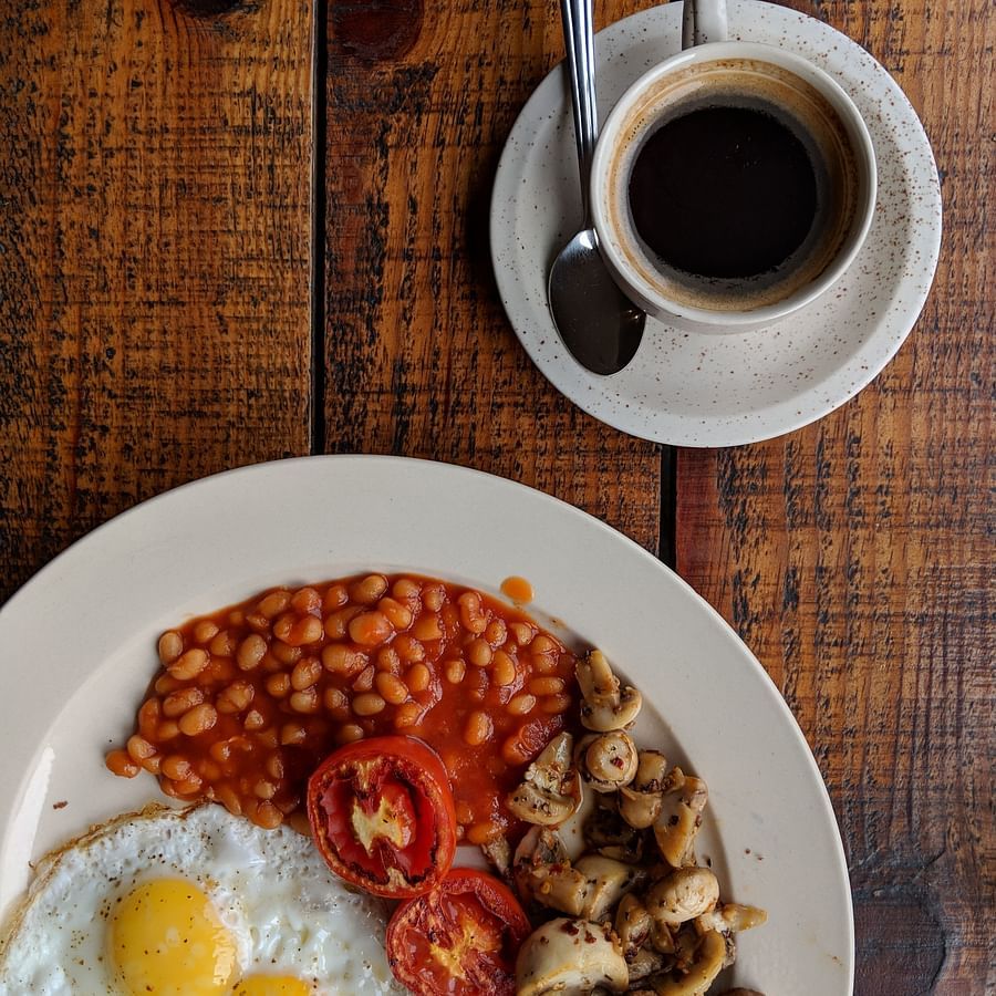 An overhead shot of English breakfast in a white plate sided with a coffee served in a white mug and saucer