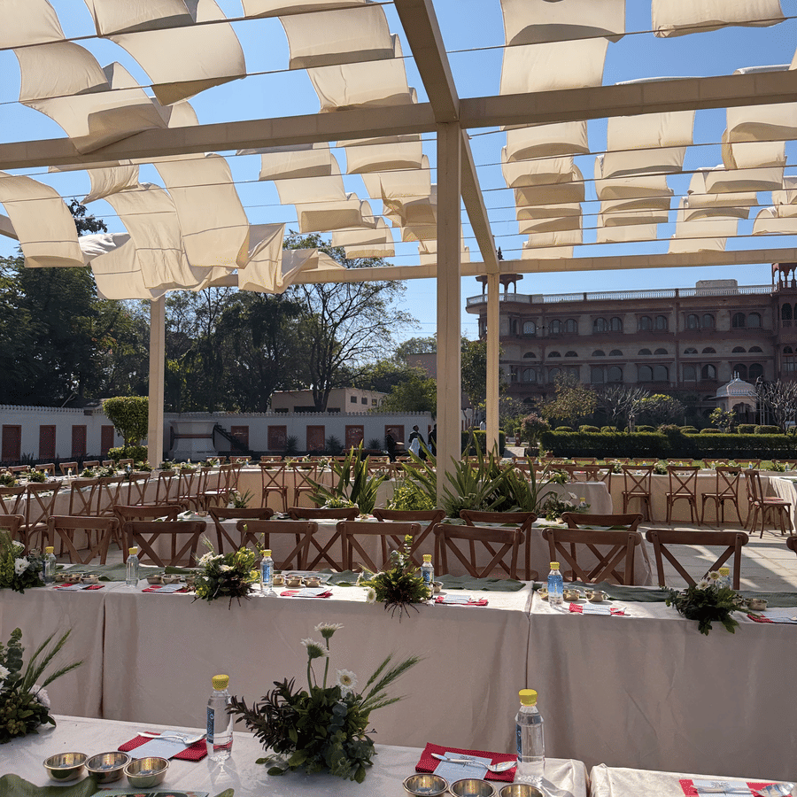 Long dining tables decorated with floral centerpieces set up on an outdoor lawn under a white wavy fabric shade canopy at Umaid Palace.