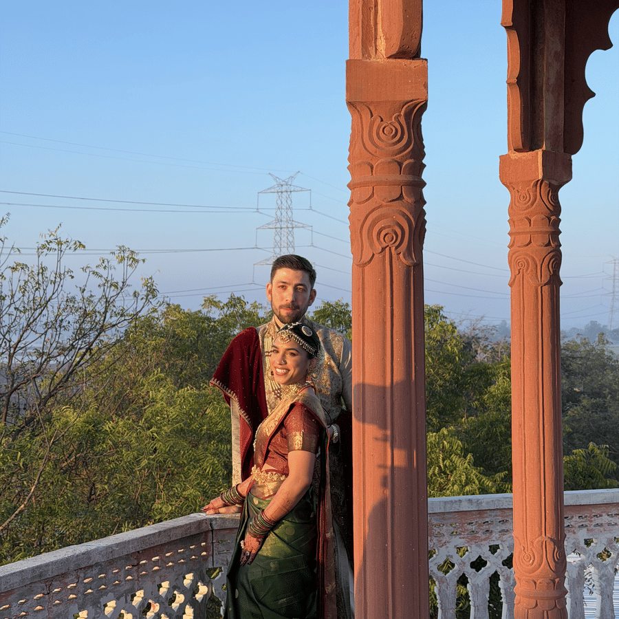 A couple in traditional attire stands by carved stone pillars on an elevated balcony overlooking trees at Umaid Palace.