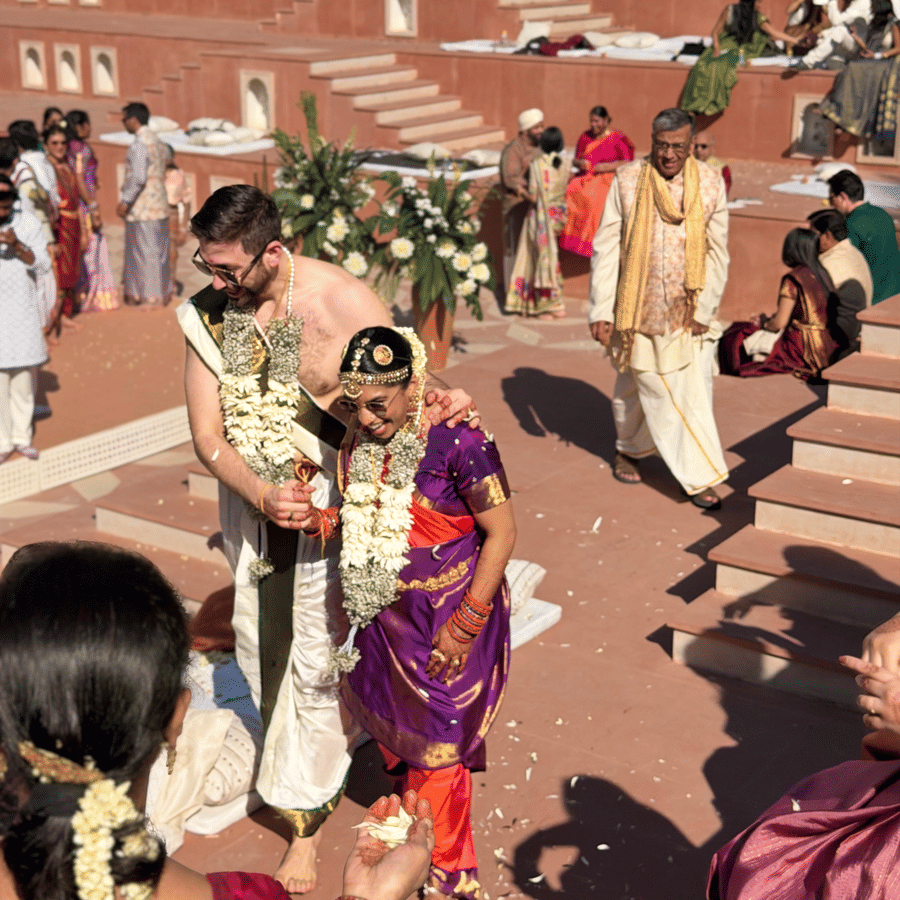 A bride and groom walk through a crowd of guests gathered on red stone steps during a daytime ceremony at Umaid Palace.