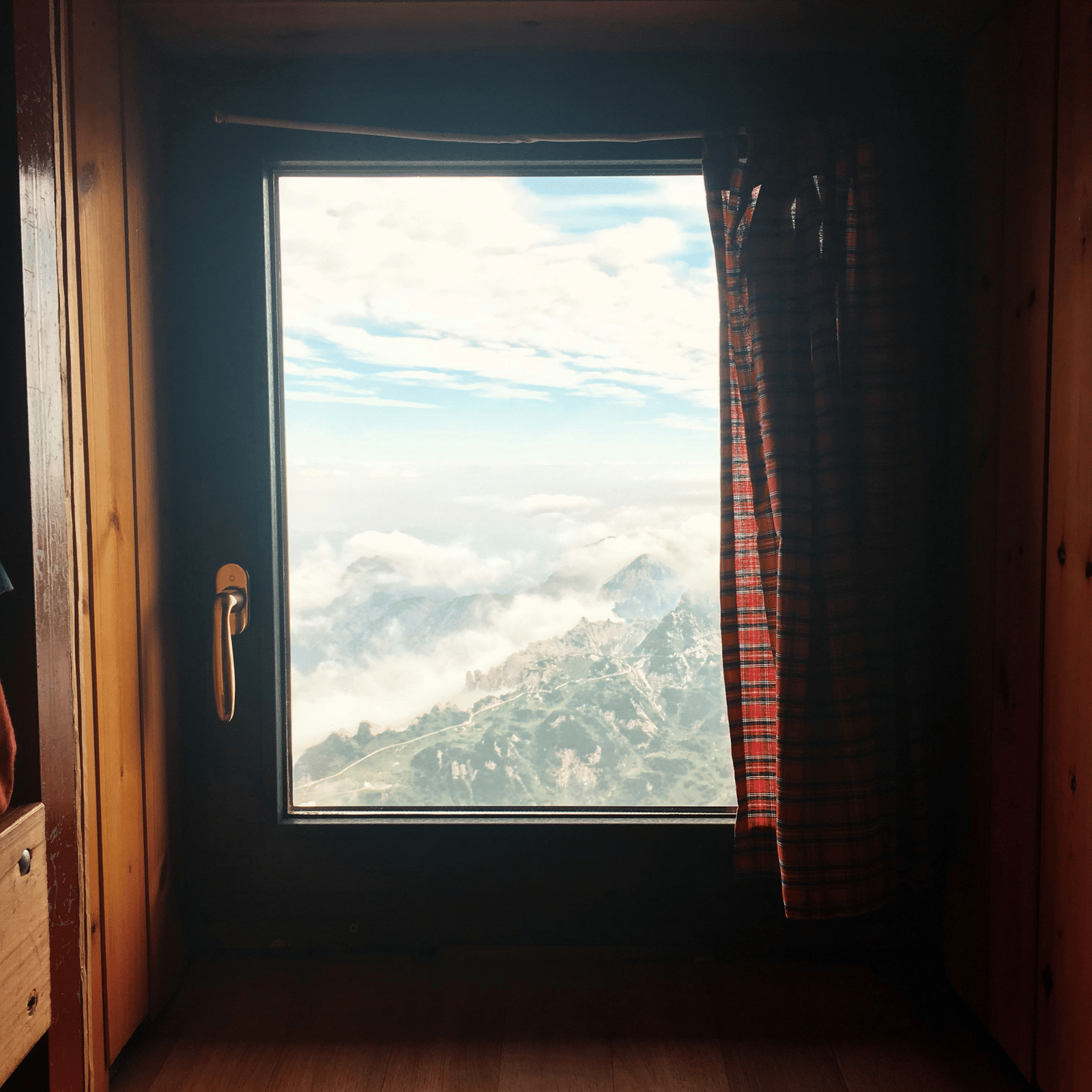 A window framed by dark wood, looking out onto a stunning vista of rolling, misty mountains under a bright, cloudy sky.