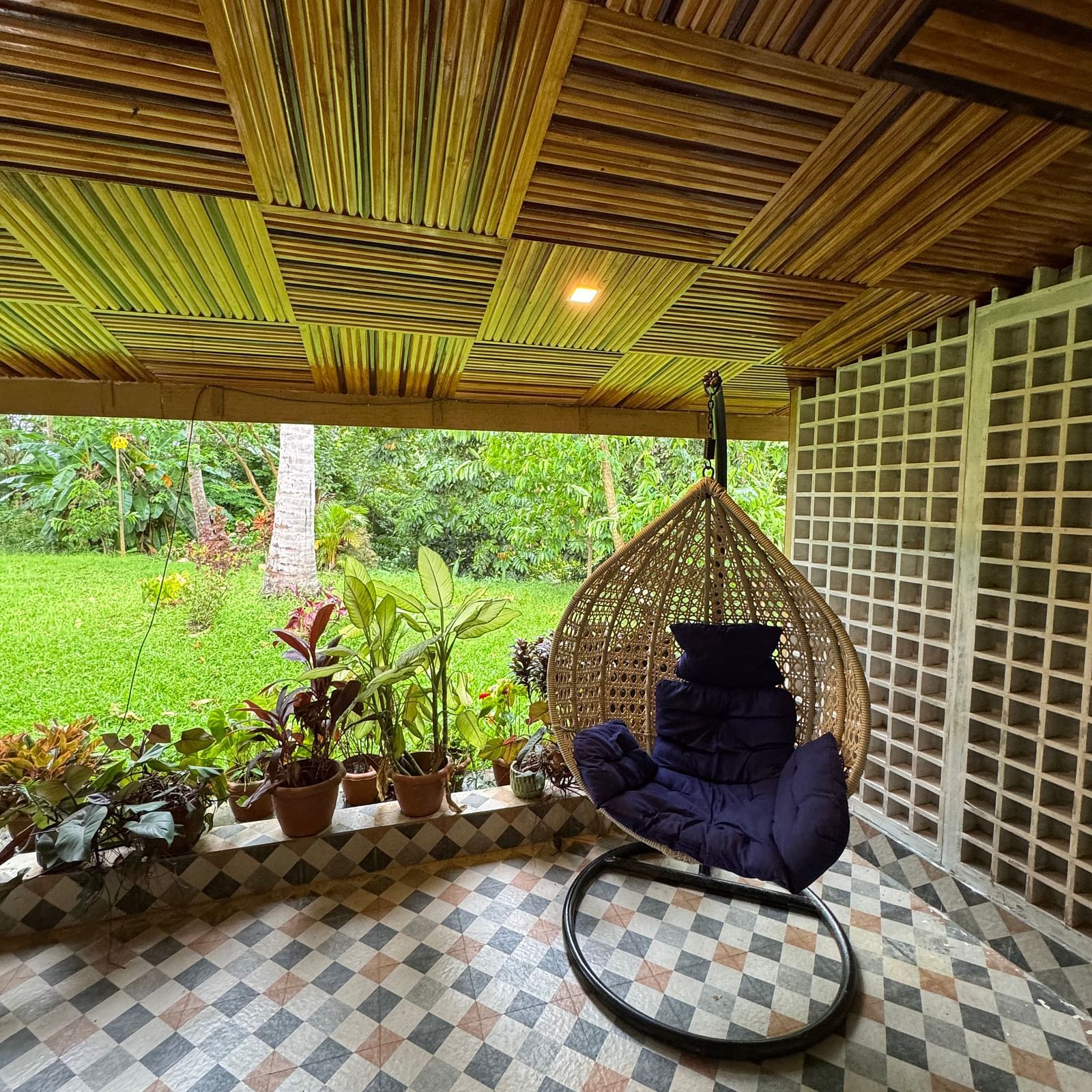 A cozy outdoor seating area with a hanging wicker chair, patterned tile floor, and a distinctive wooden ceiling.