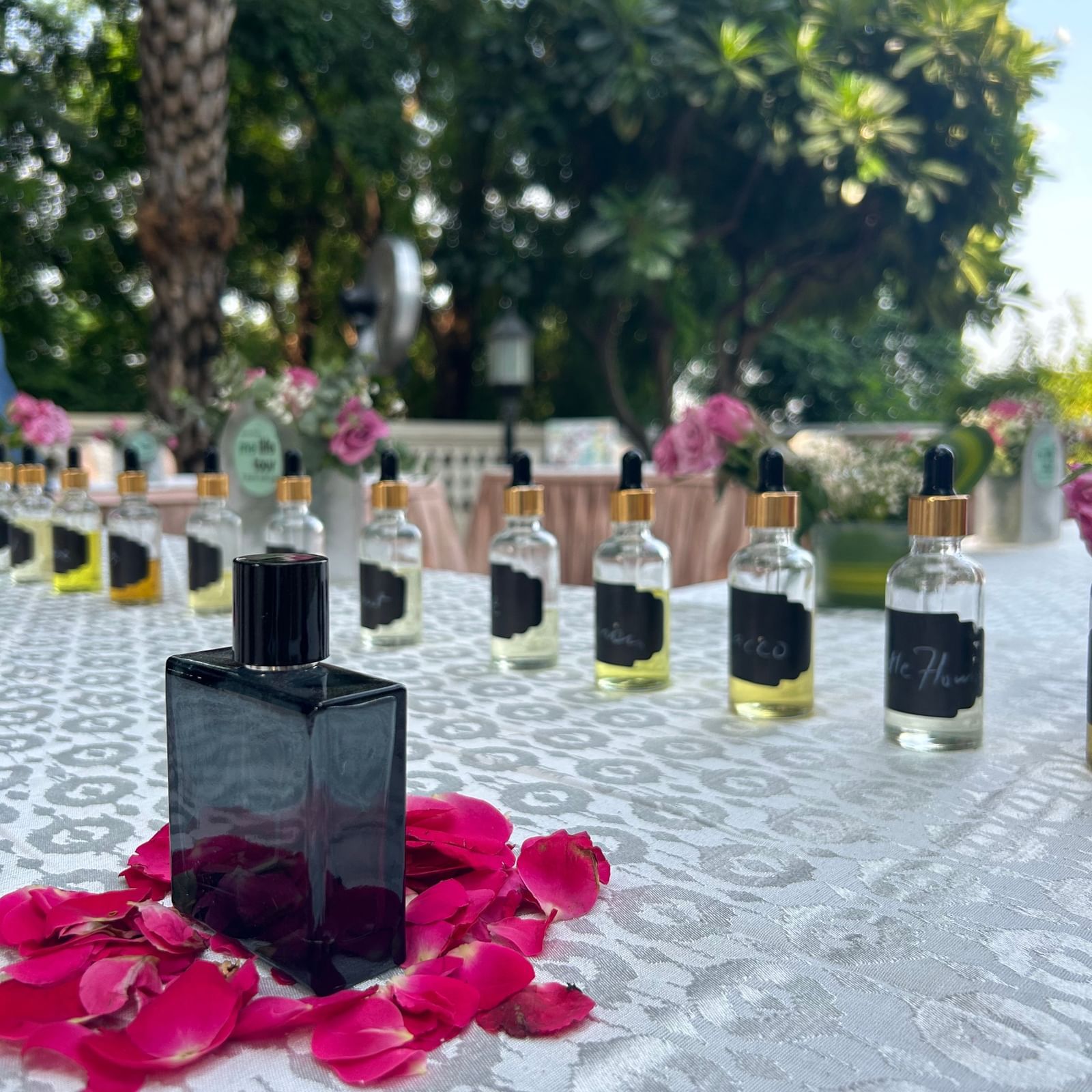 A black perfume bottle rests on pink rose petals atop a lace?covered outdoor table, with rows of clear dropper bottles and a garden backdrop behind it - Kastoor x Sawai Hospitality