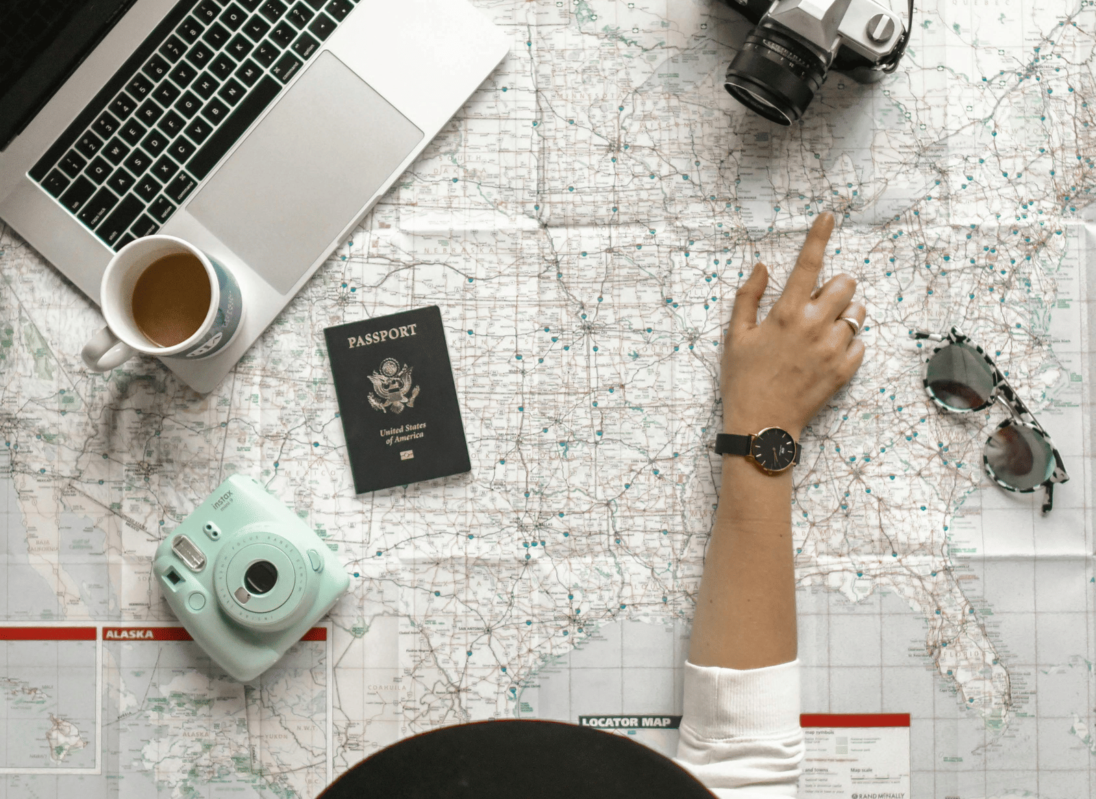 An overhead, flat-lay view of travel items—a laptop, map, sunglasses, passport, and hat—on a white surface.