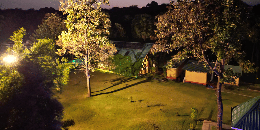 The illuminated facade of Ibex Resorts, Malampuzha (Kava Eco Camp and Caravan Park), featuring trees, and greenery at night.