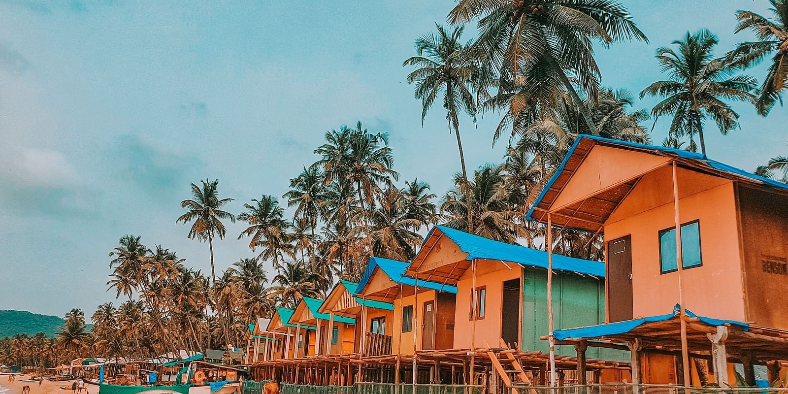 Colorful beach huts elevated on stilts, lined up along a sandy shore with palm trees in the background.