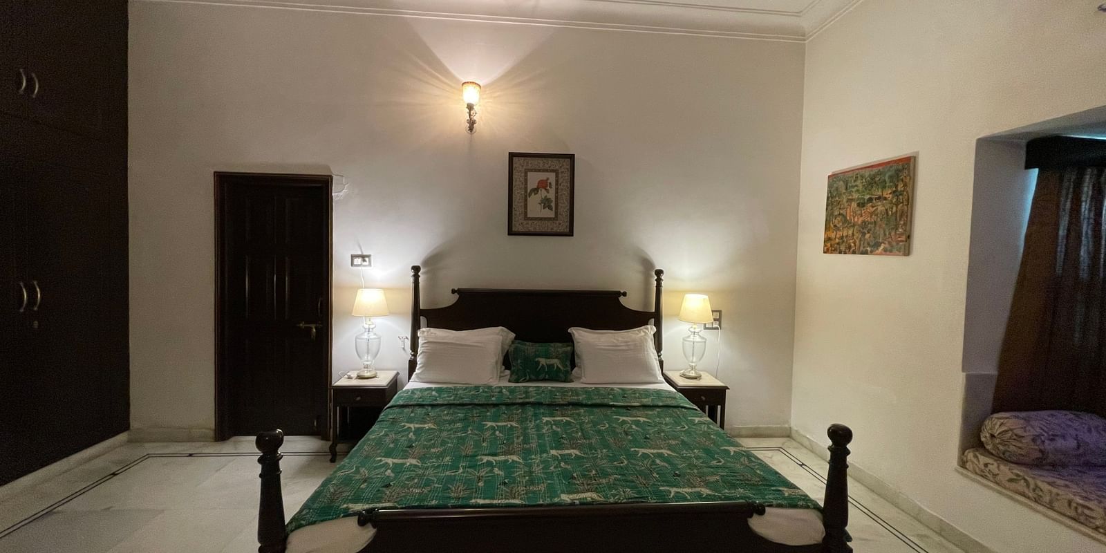 An eye-level view of the heritage room at Mahendra Niwas, showcasing the bed and a dark wooden armoire, with traditional paintings on the walls and warm lighting.