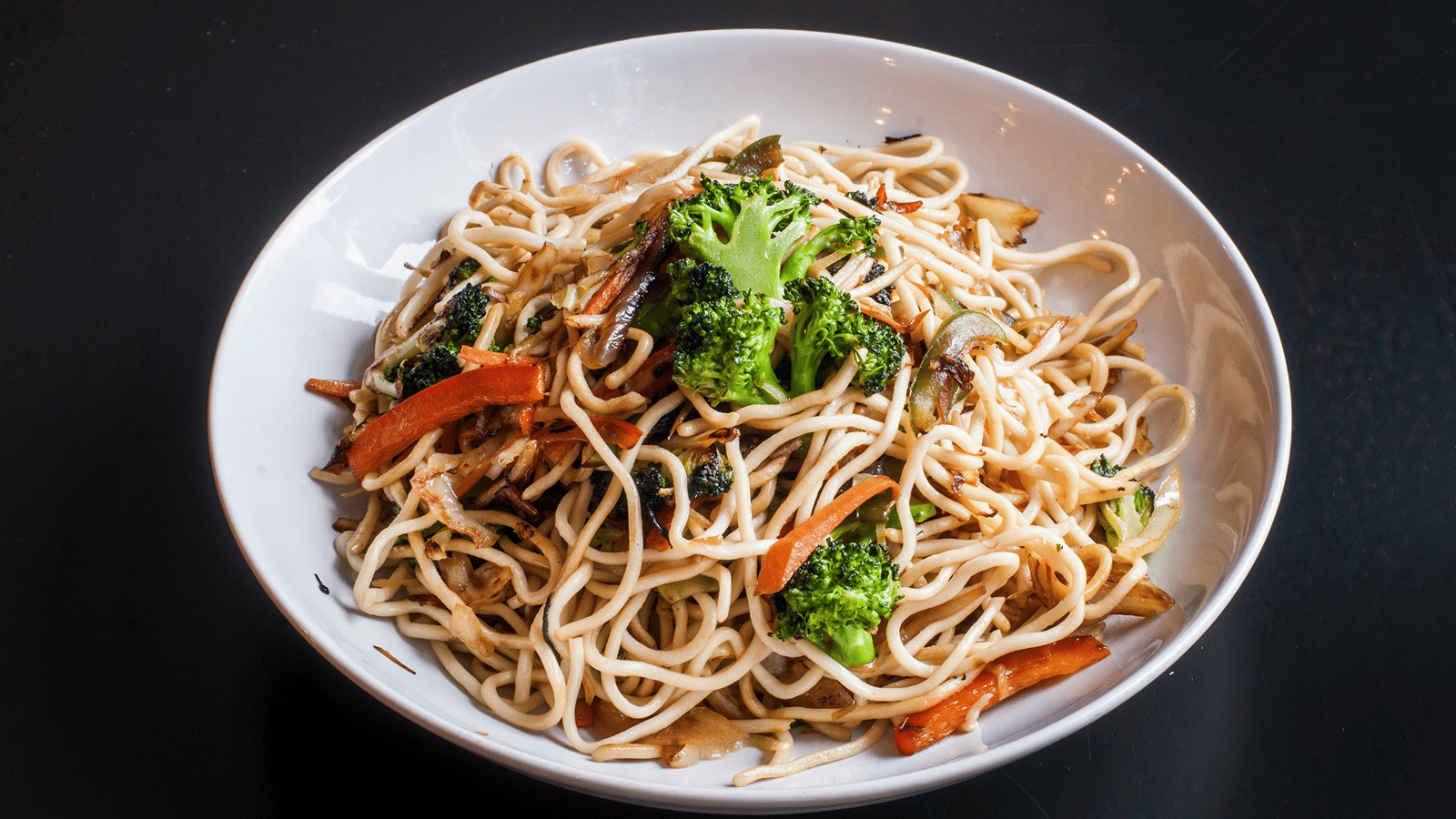 A white bowl filled with stir-fried noodles tossed with fresh broccoli florets, sliced carrots, and bell peppers on a dark background.