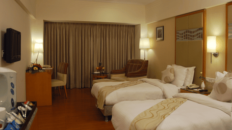 A warmly lit hotel room of Lords Plaza Ankleshwar featuring twin beds, a lounge chair and a work desk, highlighting the room's modern interiors
