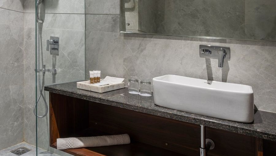 A wider view of a bathroom vanity area featuring a black granite countertop with a white basin sink, a large rectangular mirror, and a glass-enclosed shower in the background. A white towel is placed on the vanity shelf. The bathroom has grey marble walls 