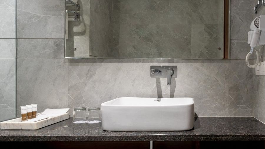A bathroom vanity with a black granite countertop, a white basin sink, and a large rectangular mirror. A white towel is neatly folded on the open wooden shelf beneath the counter. The grey marble walls and floor add a luxurious touch. - Clarks Inn Suites, 