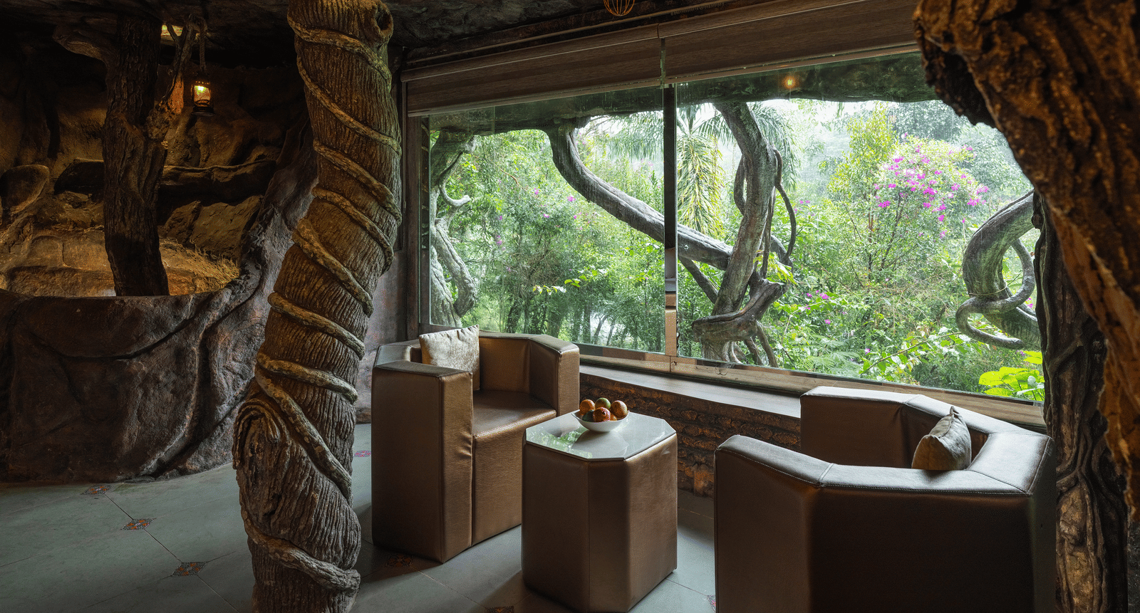 A unique bathing area in the Cave Room at Parakkat Nature Hotel and Resorts, Munnar, featuring a deep-soaking tub, a large window overlooking dense foliage, and sculpted stone-like interior walls.